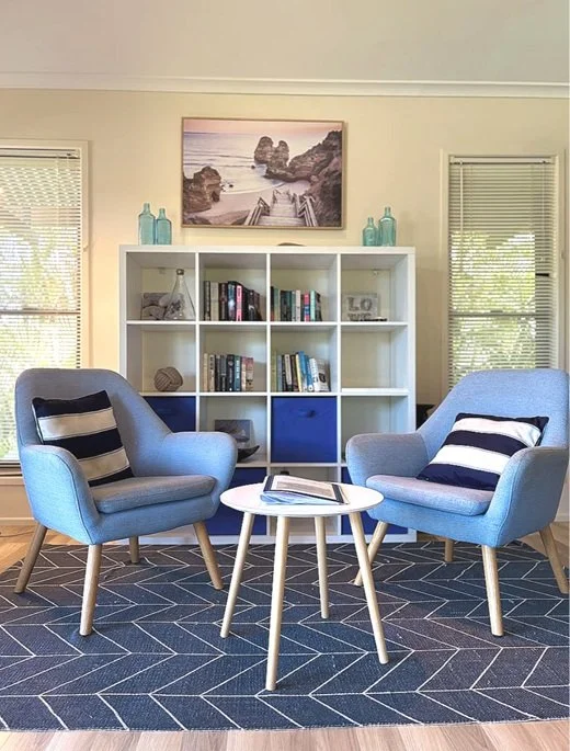 Living room with two blue armchairs, a white round coffee table, a white bookshelf with blue storage bins, a framed beach scene photograph on the wall, and windows with blinds.