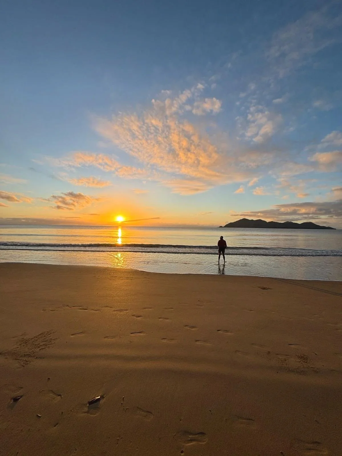 A person standing on the beach during sunrise, facing the ocean with footprints in the sand and a distant island or landmass on the horizon.