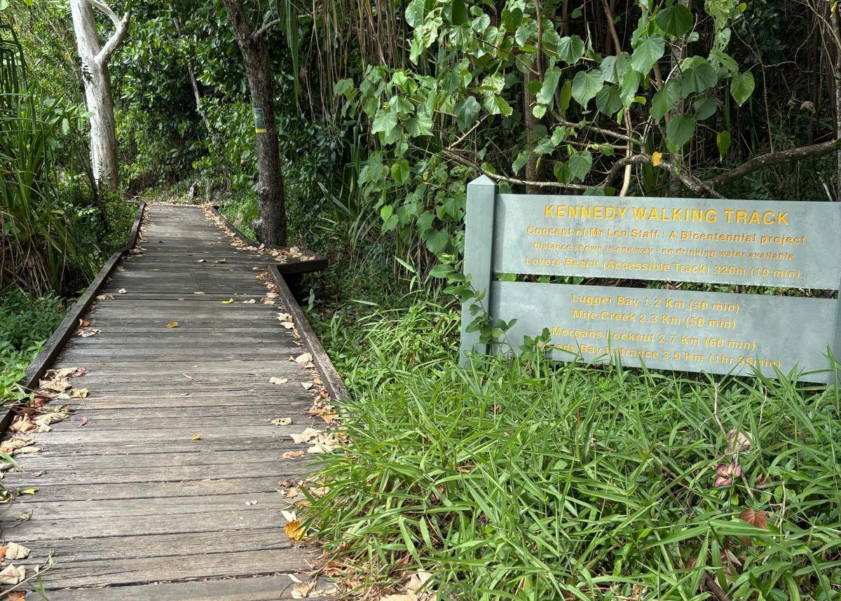 Wooden walking trail surrounded by green foliage with a sign indicating the Kennedy Walking Track and nearby destinations.