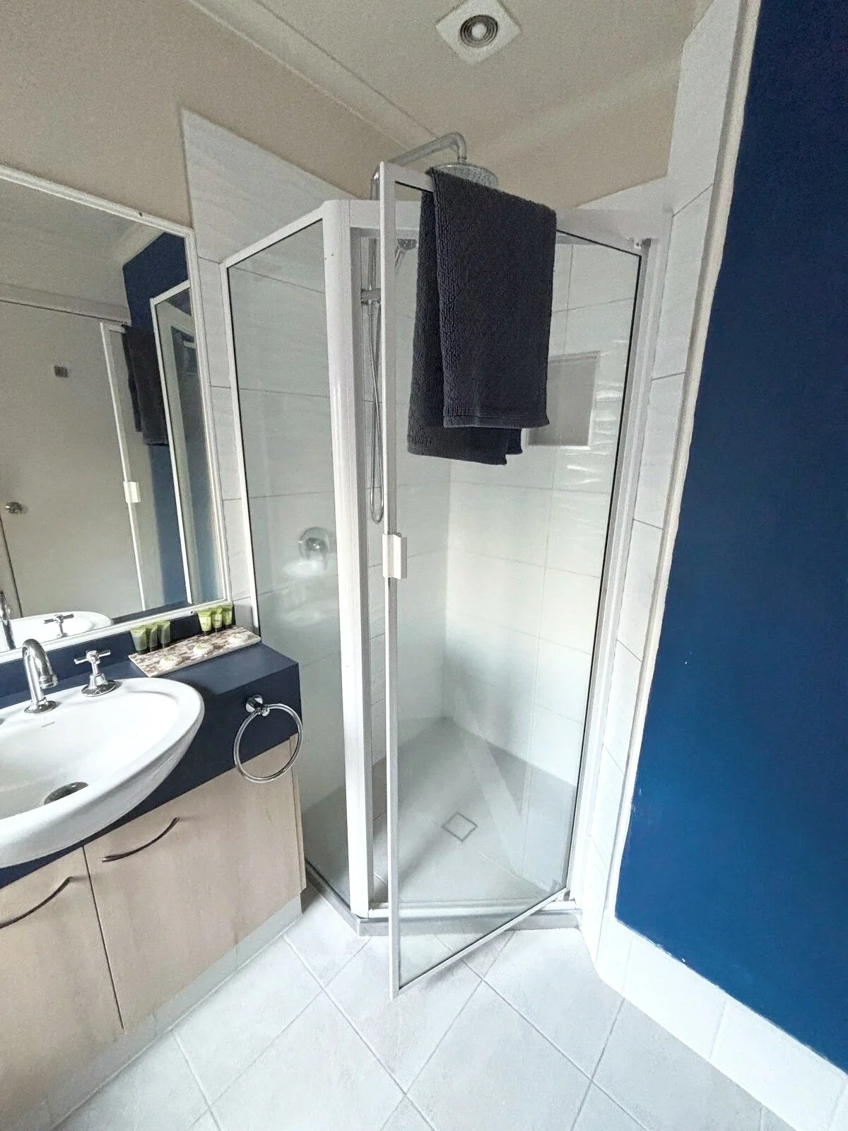 A small bathroom with a corner shower stall and a white tile wall. There is a vanity with a mirror and a small sink, with a black towel hanging from a ring below. The floor is tiled with white and black diamond patterns.