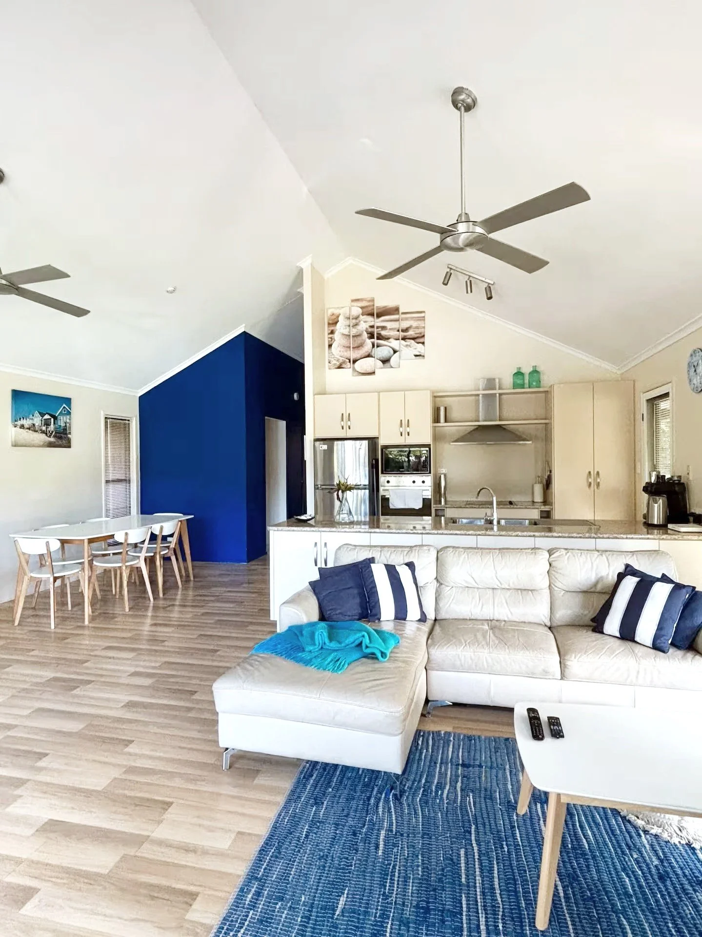 Open-concept living room and kitchen in a modern home, featuring a white sectional sofa with striped pillows, a blue area rug, a white coffee table with remote controls, an island with a sink, stainless steel appliances, a dining area with a wooden table and chairs, and a blue accent wall with wall art.