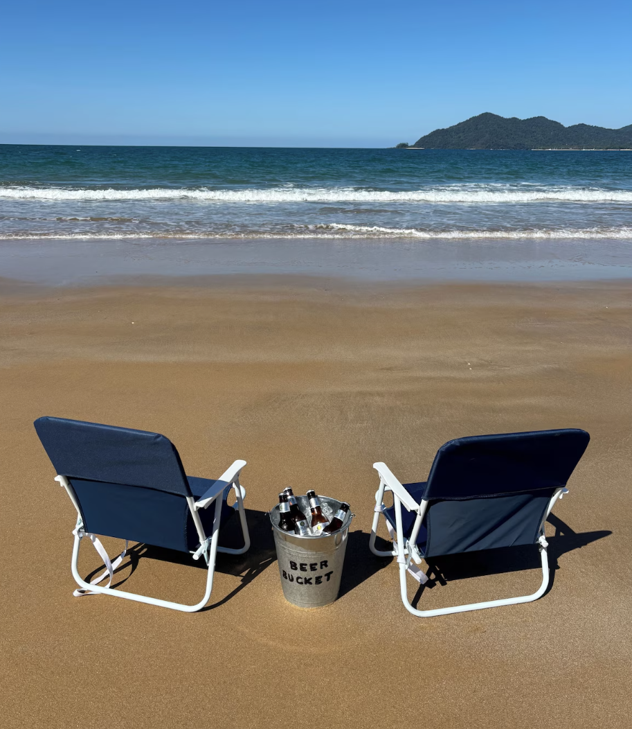 Two blue beach chairs facing the ocean on a sandy beach with a metal bucket labeled 'Beer Bucket' filled with bottles between them, and Dunk island in the distance under a clear blue sky.