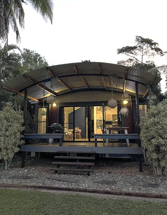 A small elevated house or cabin with a curved metal roof, surrounded by lush greenery and trees, with warm interior lights visible through glass doors, and outdoor hanging lanterns for lighting.