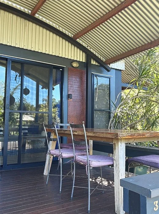 An outdoor patio with a wooden table and purple cushioned chairs, surrounded by greenery, with a corrugated metal roof overhead and sliding glass doors leading into a house.