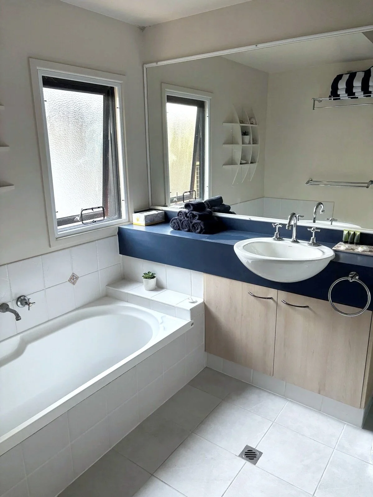 Bathroom with a bathtub, double windows, a sink with a mirror, shelves with shells, and blue towels.