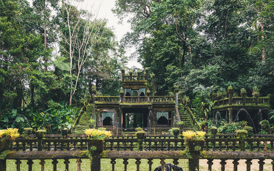 Moss-covered stone structure in a lush, green tropical garden with multiple levels, staircases, fountains, and potted plants.
