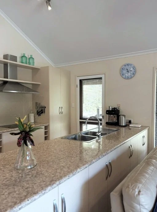 Kitchen with beige cabinets, granite countertop, a vase with flowers, a sink, coffee machine, kettle, and wall clock showing 11:11.