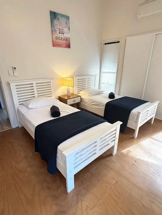 A bedroom with two single beds, each with a white headboard, white bedding, and a rolled black towel on top. There is a nightstand with a lamp between the beds, a window with blinds, and a framed picture on the wall reading 'Dreams as Big as the Ocean.'