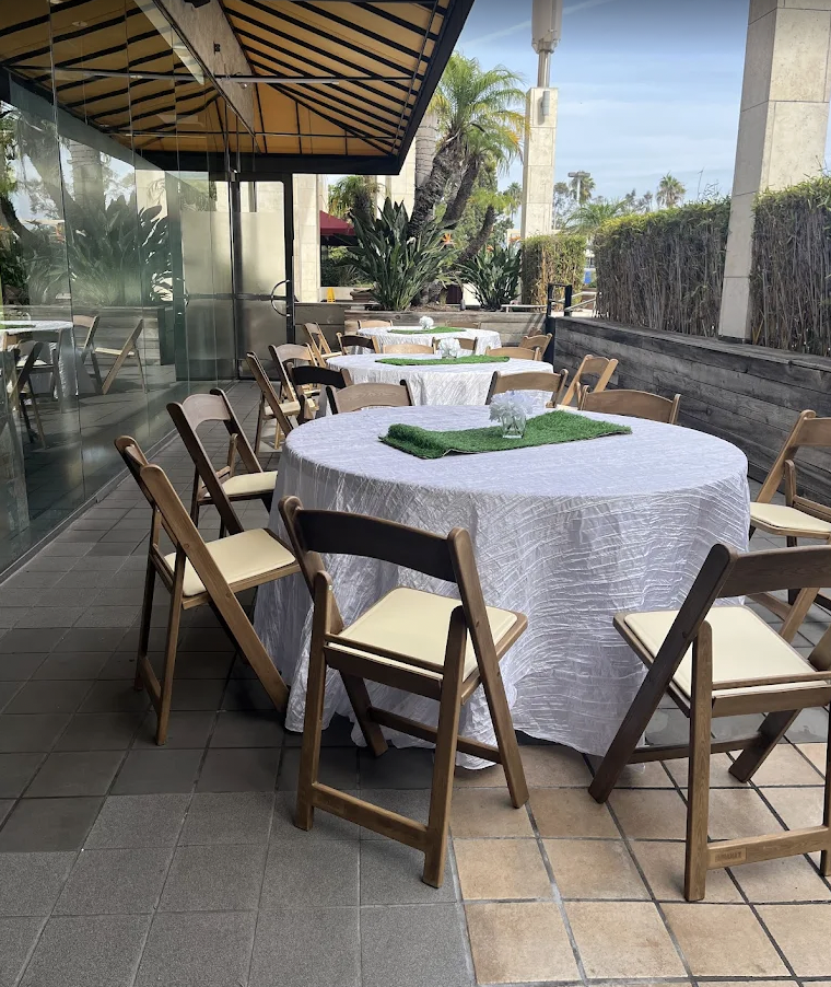 Outdoor restaurant patio with several round tables covered with white tablecloths, surrounded by wooden chairs with cream-colored cushions, and decorated with small glass vases and green table runners.