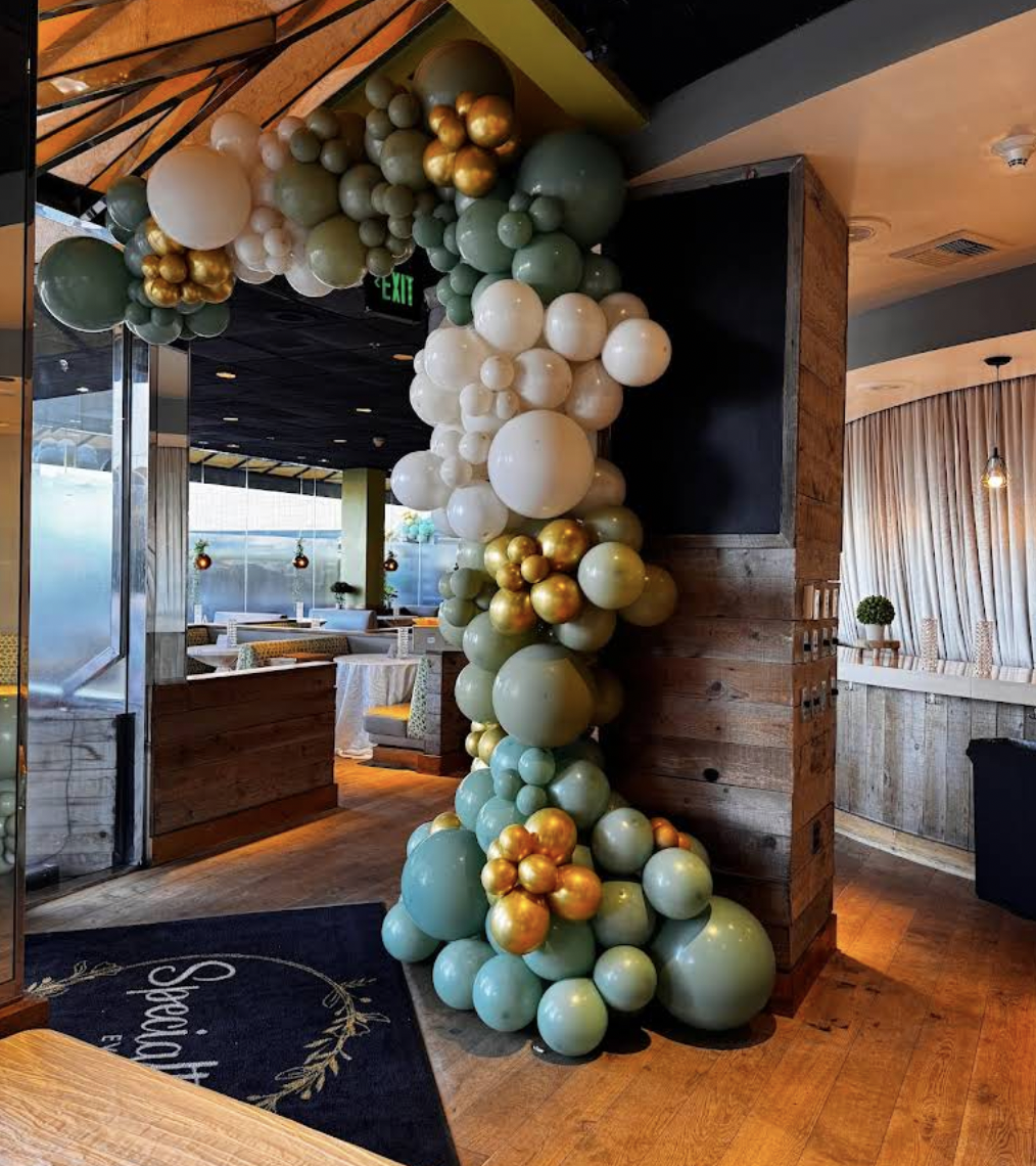 Decorative balloon arrangement in shades of green, white, gold, and beige cascading from ceiling to floor inside a restaurant.