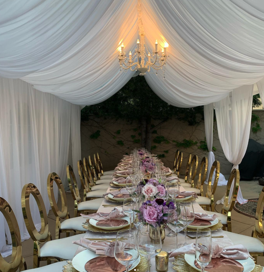 Elegant outdoor dining setup with a long table, white and pink floral centerpiece, wine glasses, pink napkins, gold-rimmed plates, and a white draped canopy with a chandelier overhead.