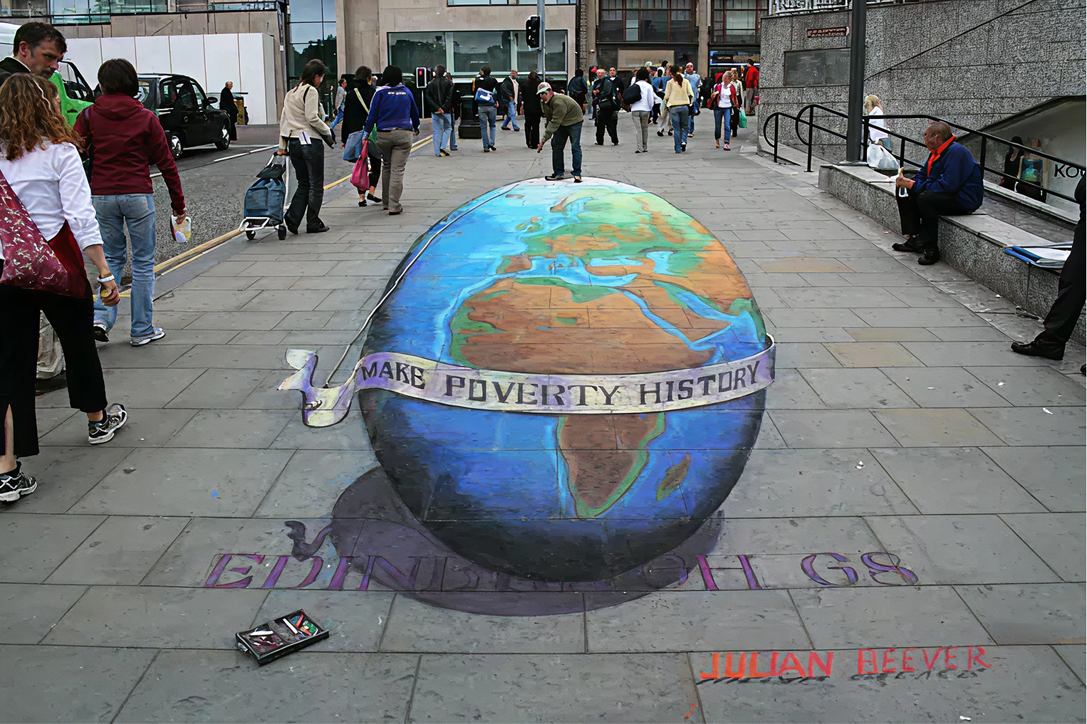 Street art of a 3D globe on pavement with banners reading 'Make Poverty History' and 'Ending Poverty'. Surrounding people walking, sitting, and observing.