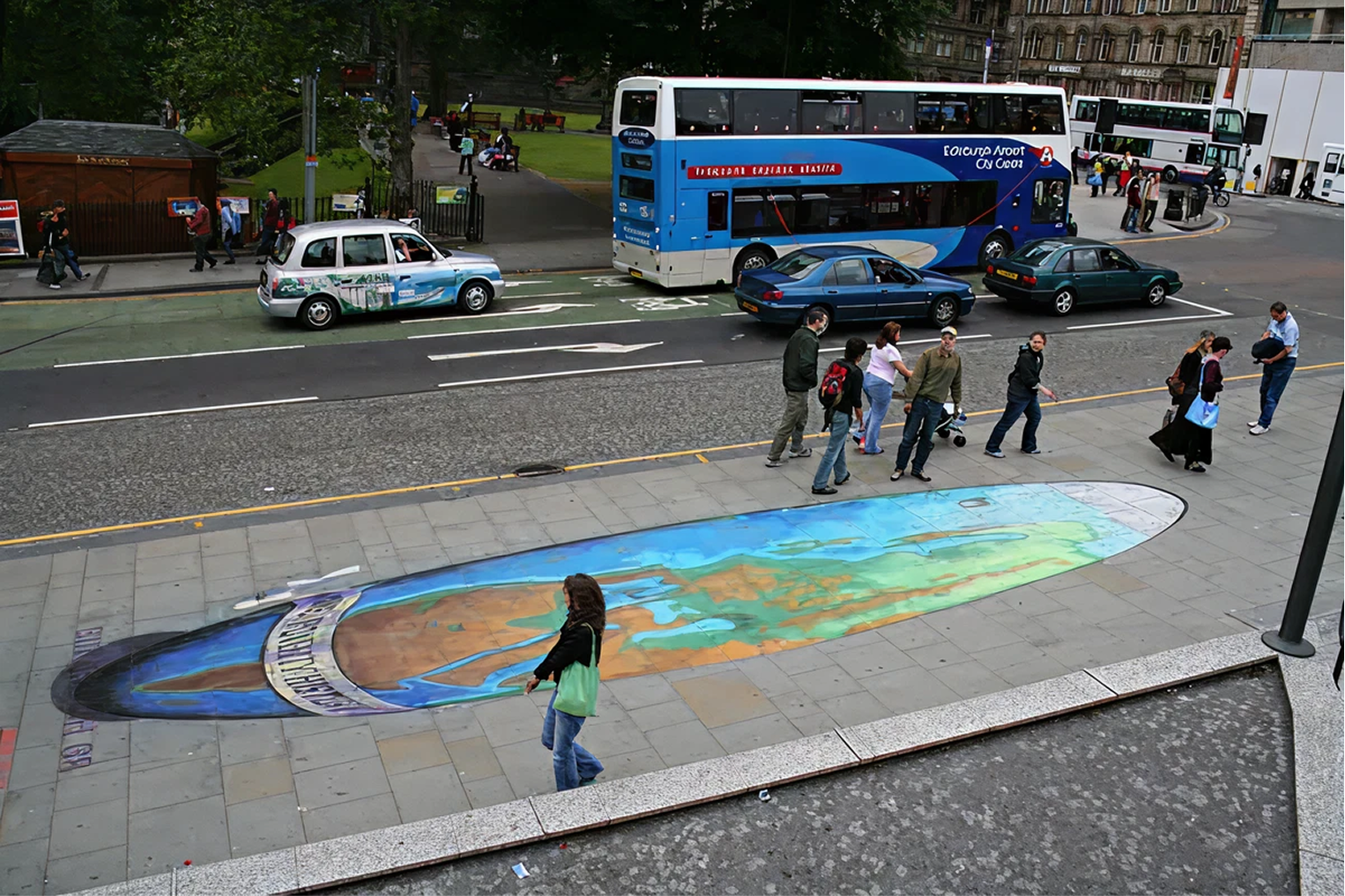 Street scene with people walking on sidewalk, parked cars, double-decker bus, and a colorful rainbow-themed street art piece on the pavement.