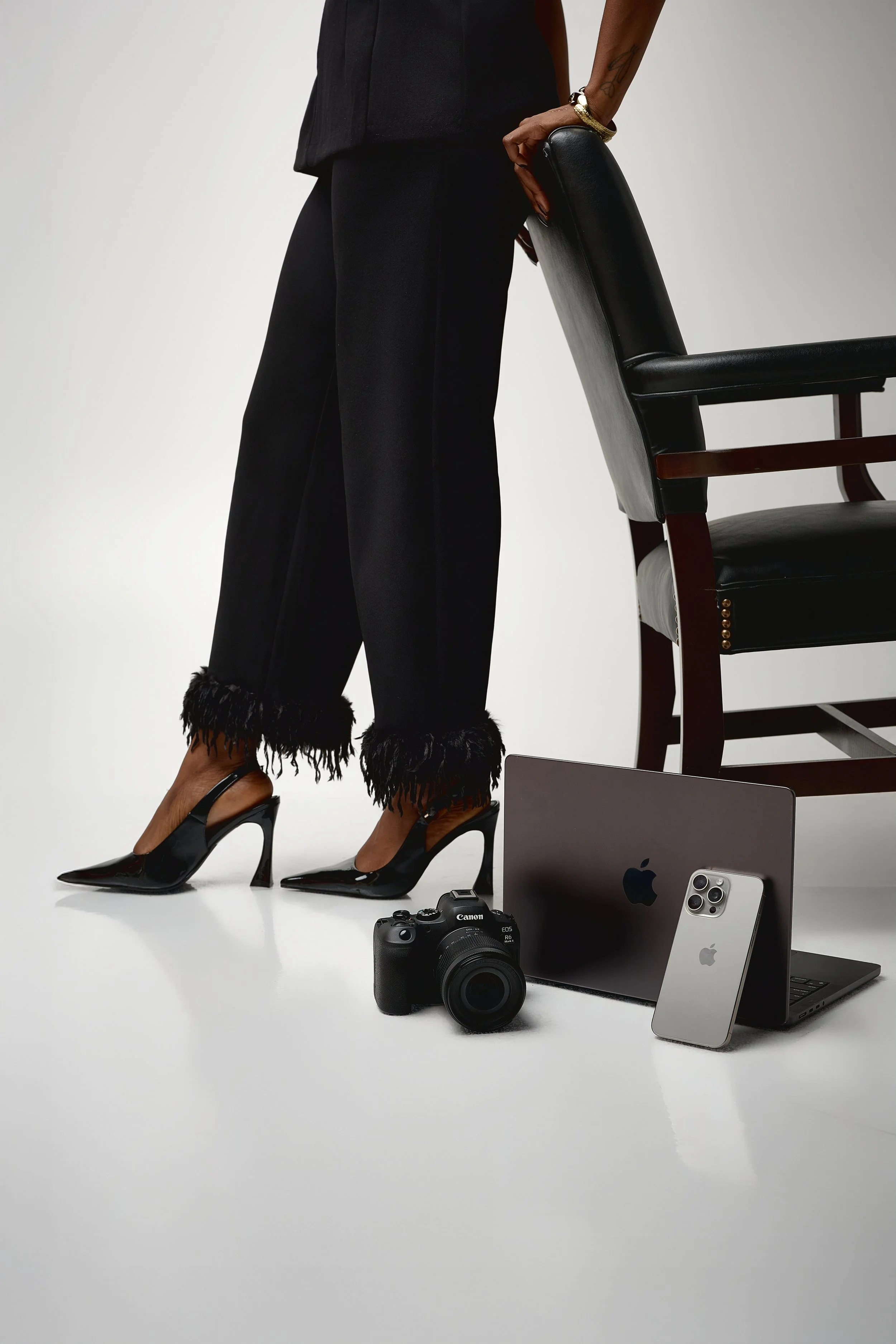 Close up photo of a woman's legs with black heels on holding on to the back of a chair while a laptop, phone, and camera surround the chair legs.