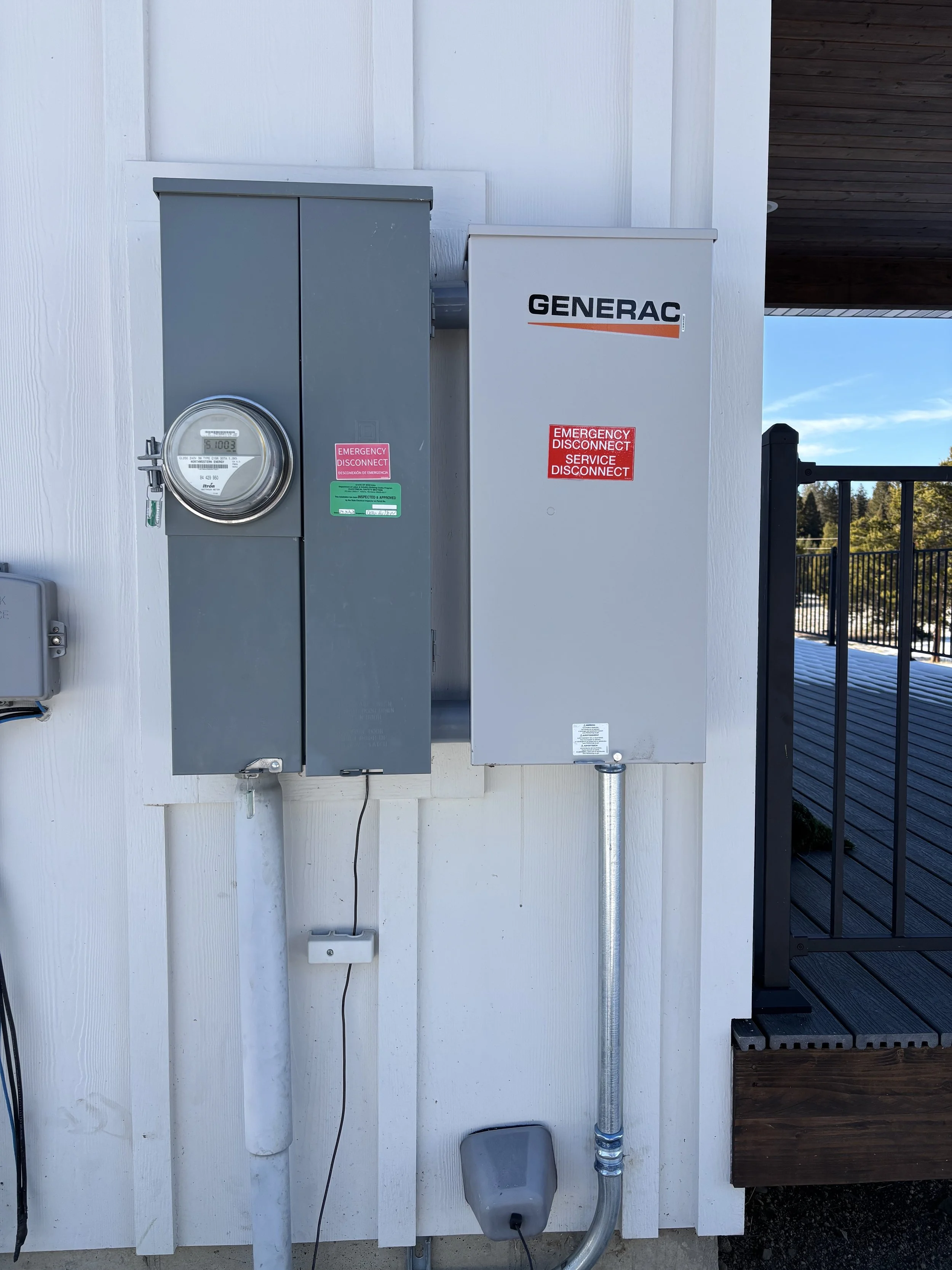 Utility boxes and electrical meter mounted on a white exterior wall outside a house, with a black railing and wooden deck on the right side.