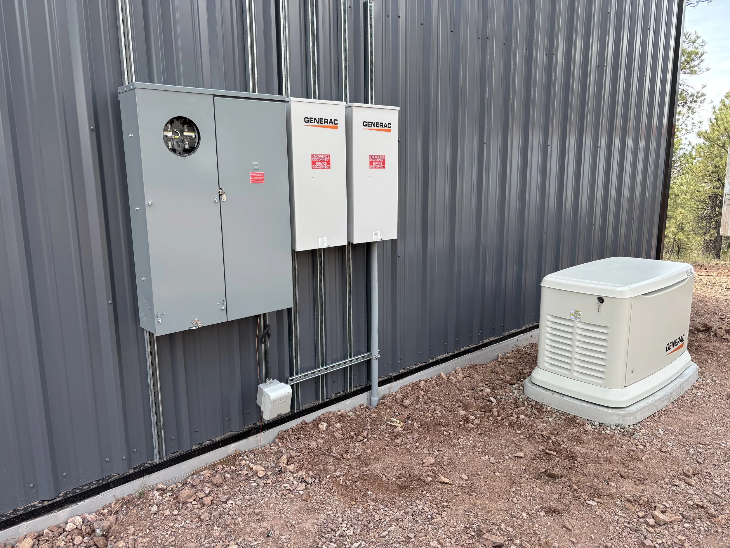 Electrical panels labeled with Generac and emergency service sticker, installed on a metal wall outside, next to an air conditioning unit on rocky ground with trees in the background.