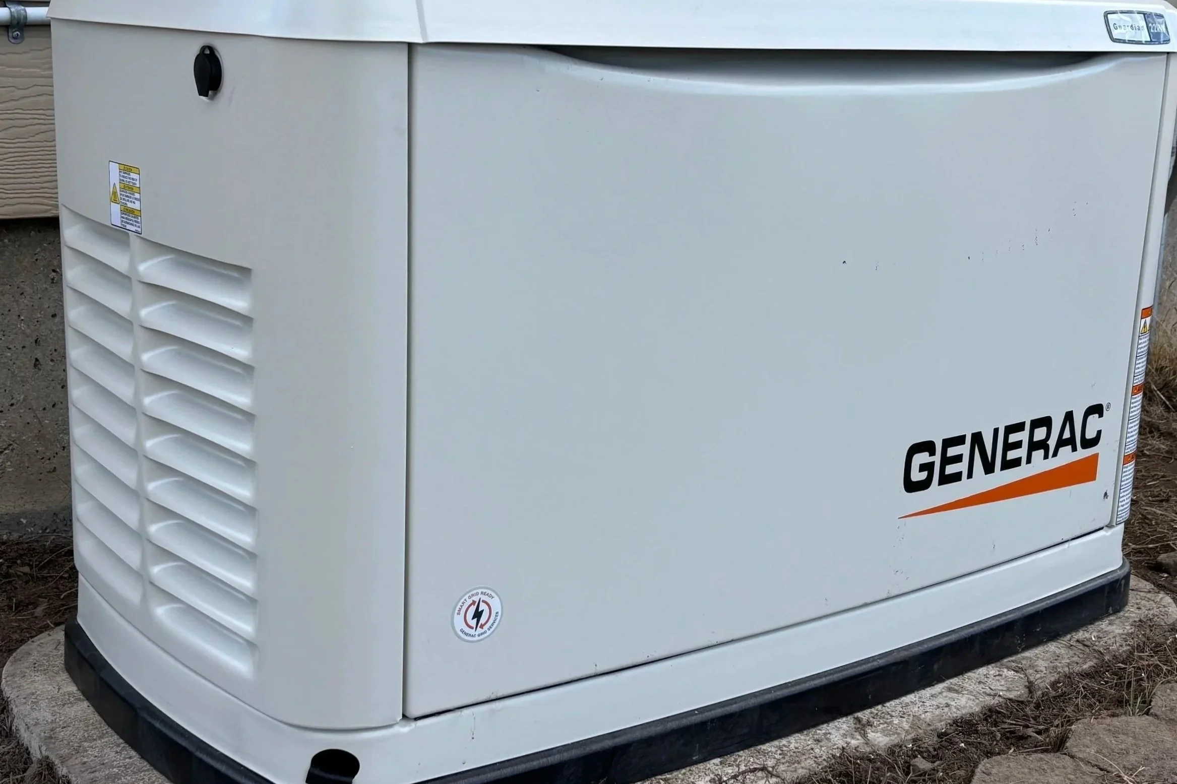 A white Generac portable generator with vented sides and a black base, placed on a concrete pad outdoors. The generator has warning stickers and a power output label on the top right corner.
