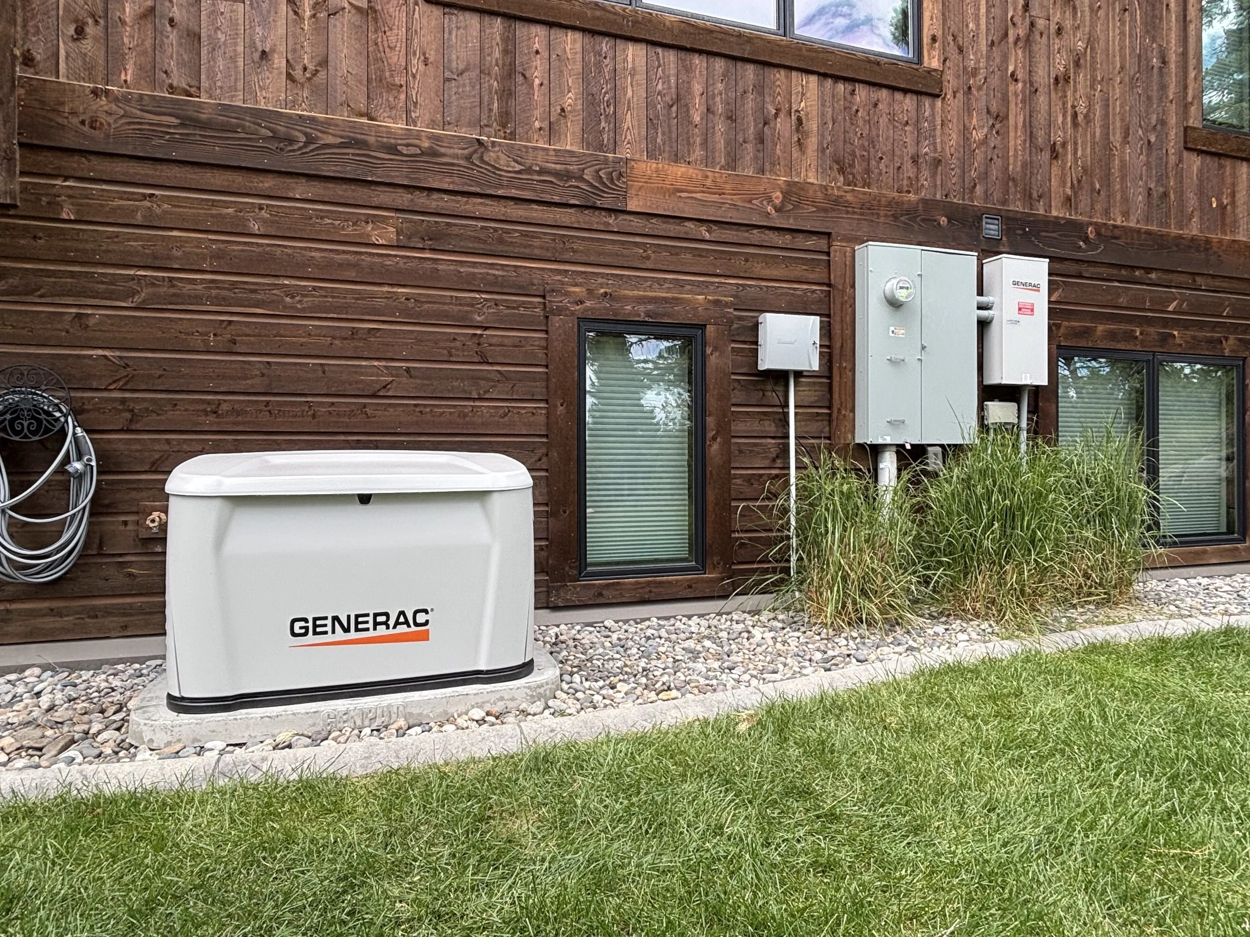 The image shows the exterior side of a wooden house with horizontal and vertical wooden planks. There is a window, a utility box, and an electrical transformer outside. Green grass and some plants grow along the base of the house, and there is gravel on the ground.