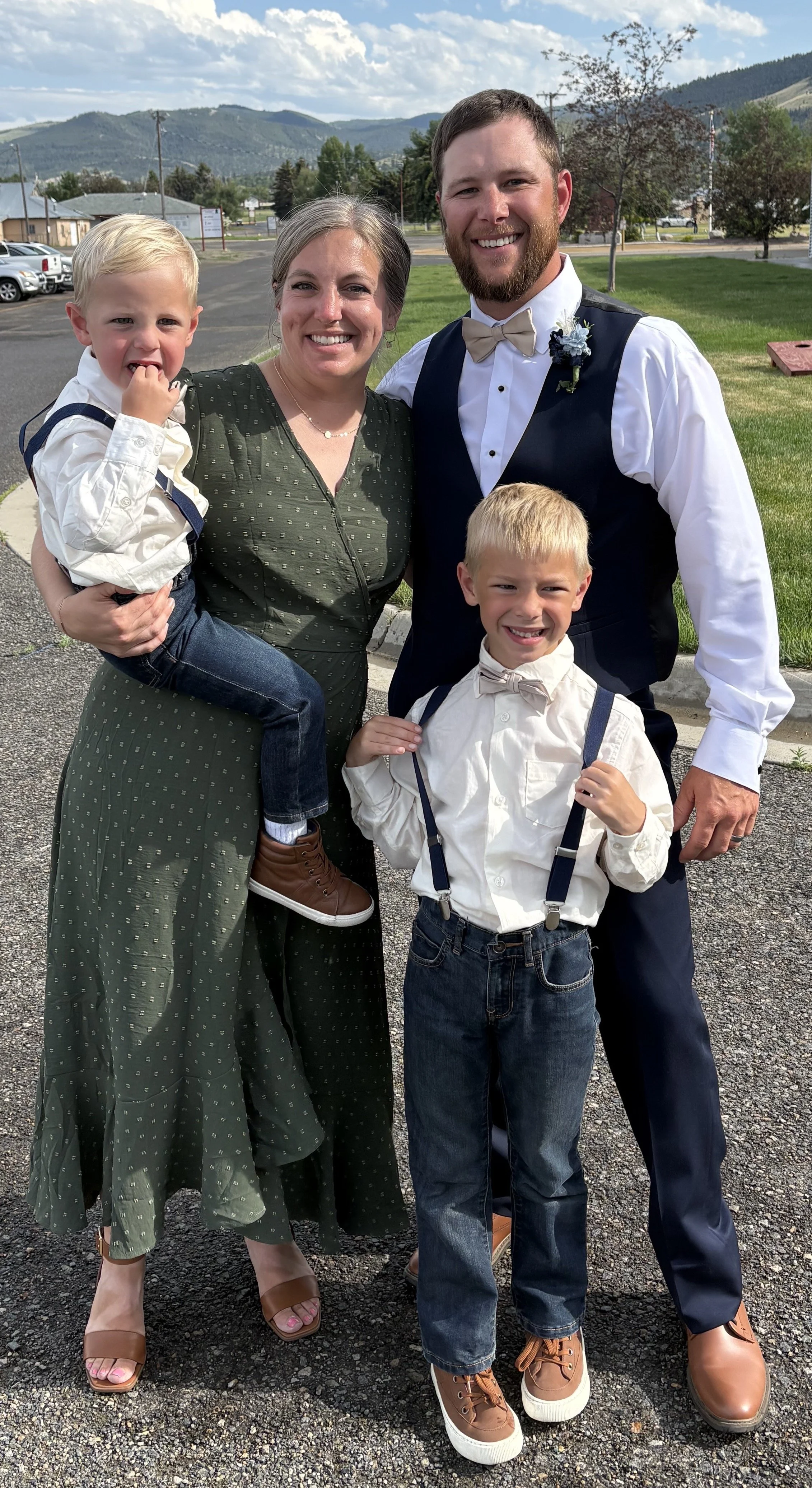 A smiling woman in a green dress holding a young boy with blond hair and wearing a white shirt. Next to them, a man with a beard in a formal suit with a bow tie and boutonniere, and another young boy with blond hair, a white shirt, suspenders, and sneakers. They are outdoors with a green lawn, trees, houses, and mountains in the background.