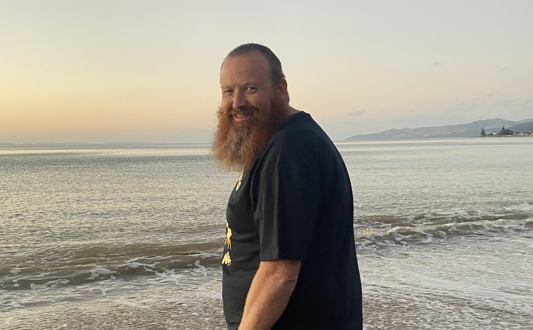 A man with a red beard and long hair, smiling, standing on a beach at sunset.