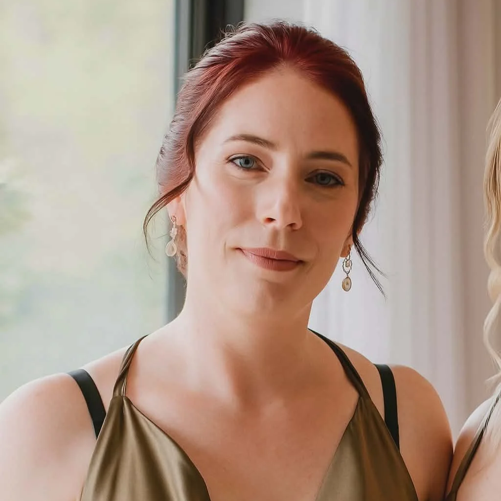 Close-up of a woman with red hair, wearing earrings and a gold-colored top, smiling gently indoors near a window