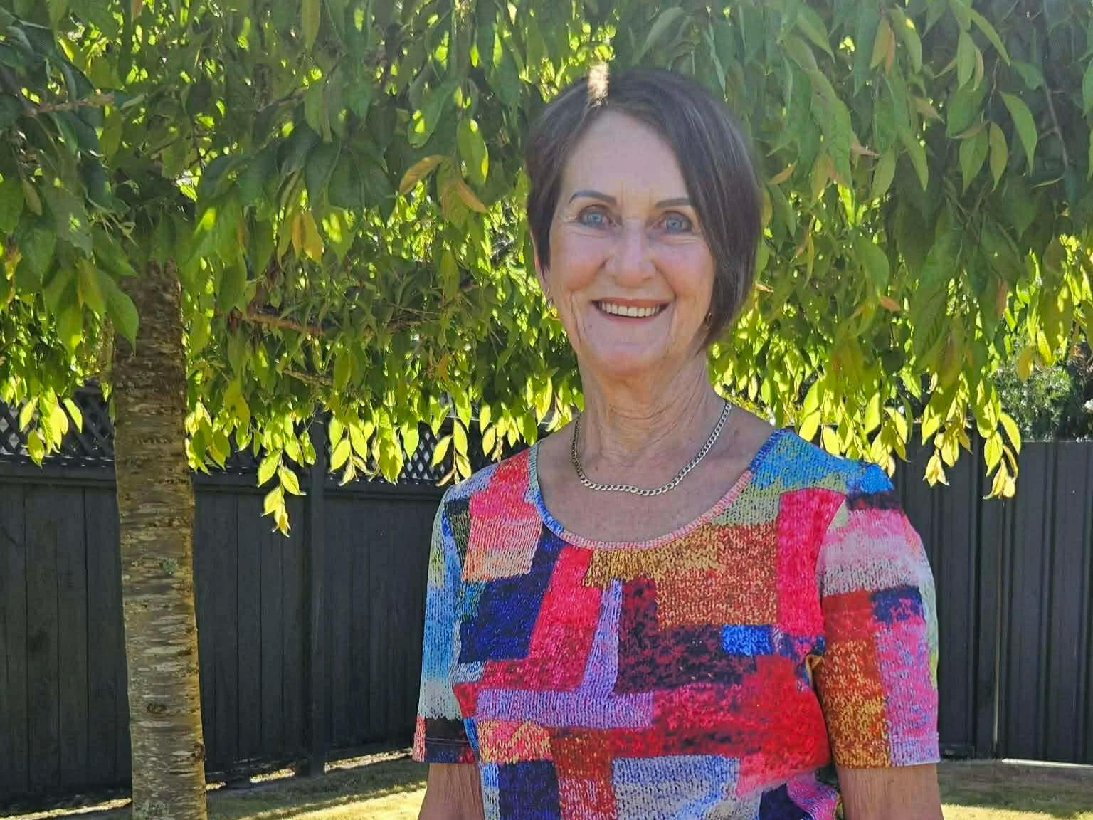 A smiling woman with short dark hair standing in front of a leafy green tree and black fence, wearing a colorful patchwork short-sleeve top and a gold necklace.