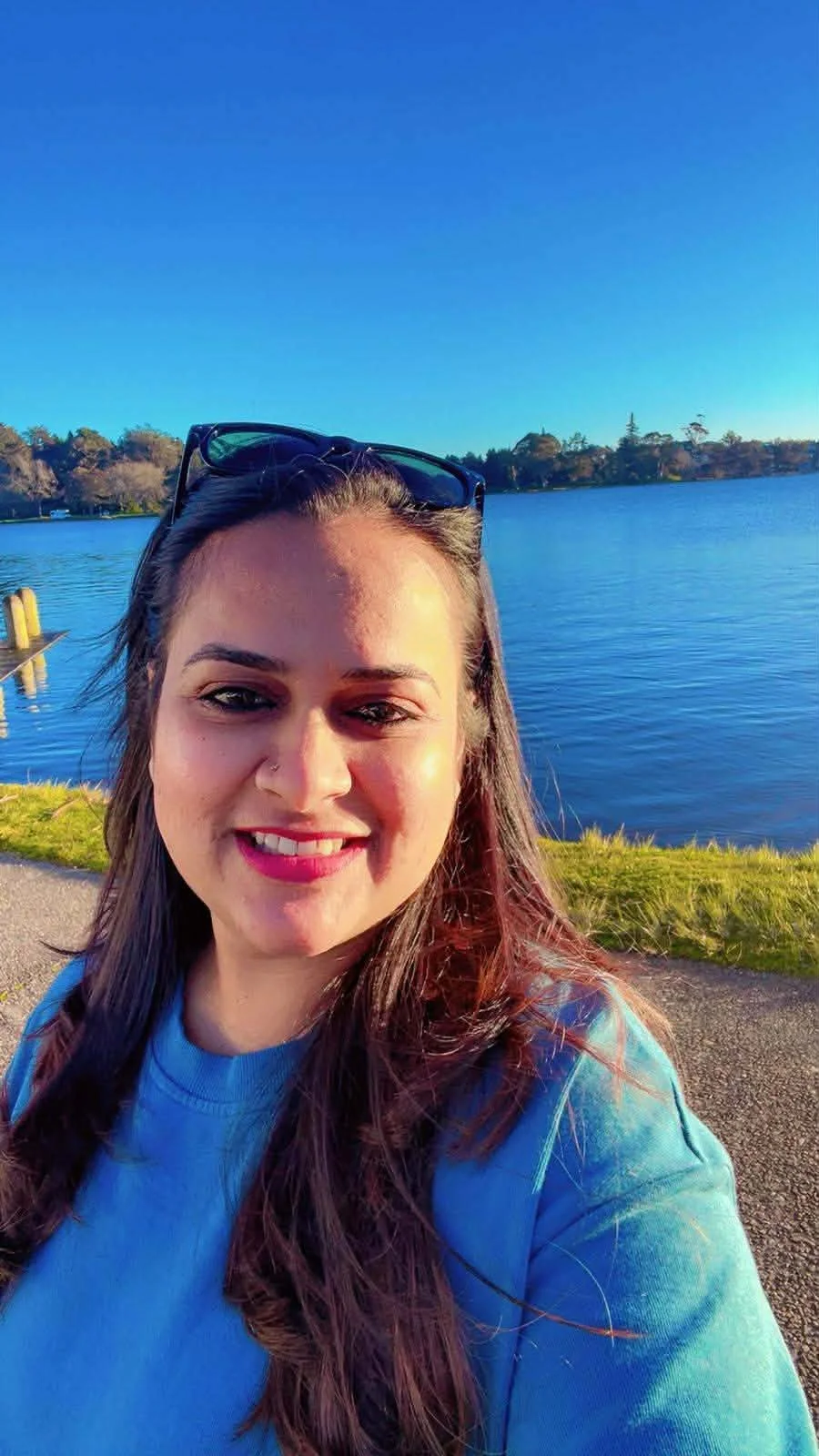 A young woman taking a selfie outdoors near a body of water on a sunny day, wearing a blue shirt and sunglasses over her head, with trees and a clear blue sky in the background.