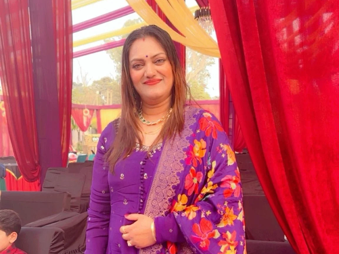 A woman in a purple saree with a floral pattern, standing inside a decorated tent with red drapes and yellow accents, smiling at the camera.