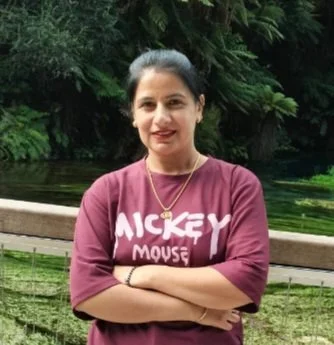 A woman with dark hair standing outdoors in front of trees, wearing a maroon Mickey Mouse shirt and a gold necklace.
