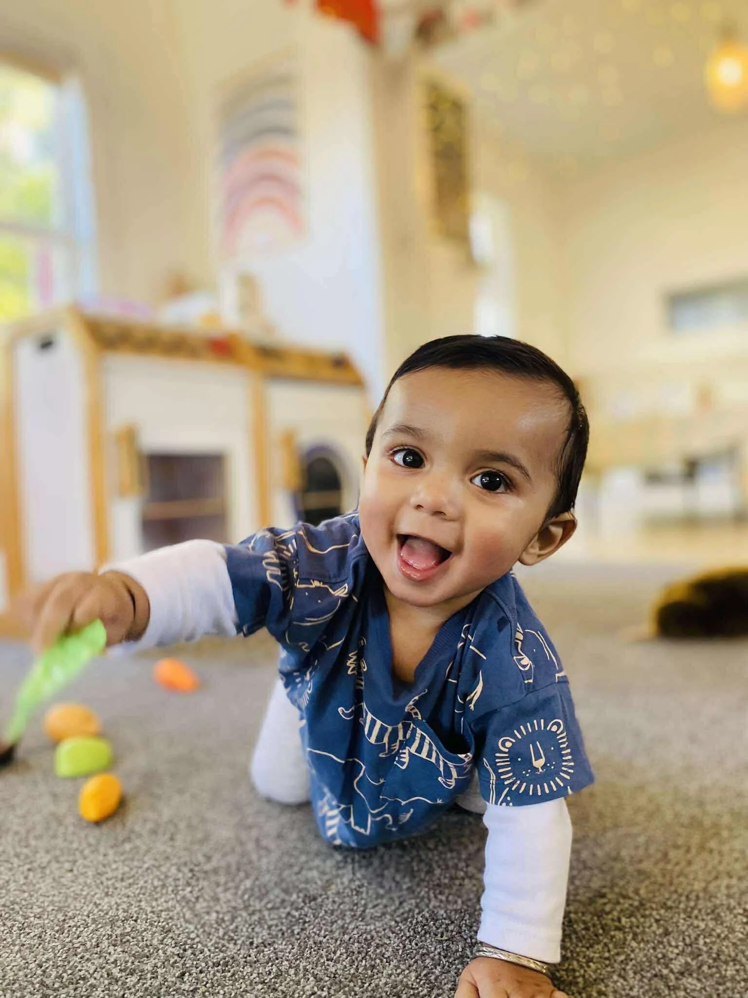 A smiling baby crawling on a carpeted floor in a playroom, reaching towards a toy, with colorful decorations and furniture in the background.