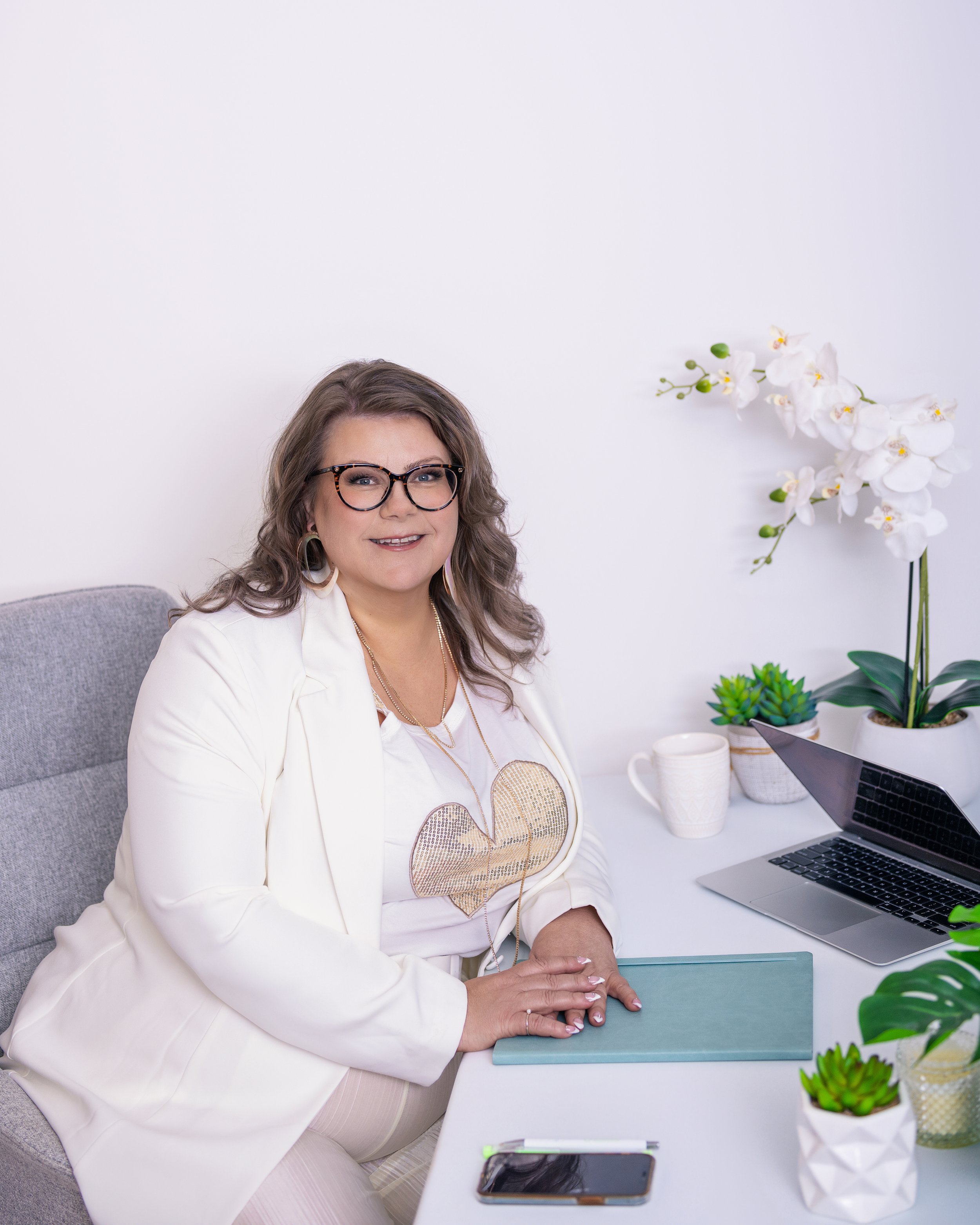 A woman with glasses and wavy hair sitting at a desk with a laptop, plants, a mug, a plant, and a tablet in an office with white walls.