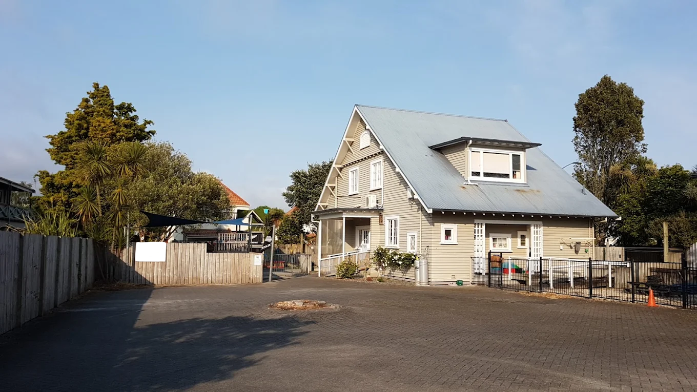 A house with a gray roof, beige siding, and white trim, surrounded by a wooden fence and trees, under a clear blue sky.