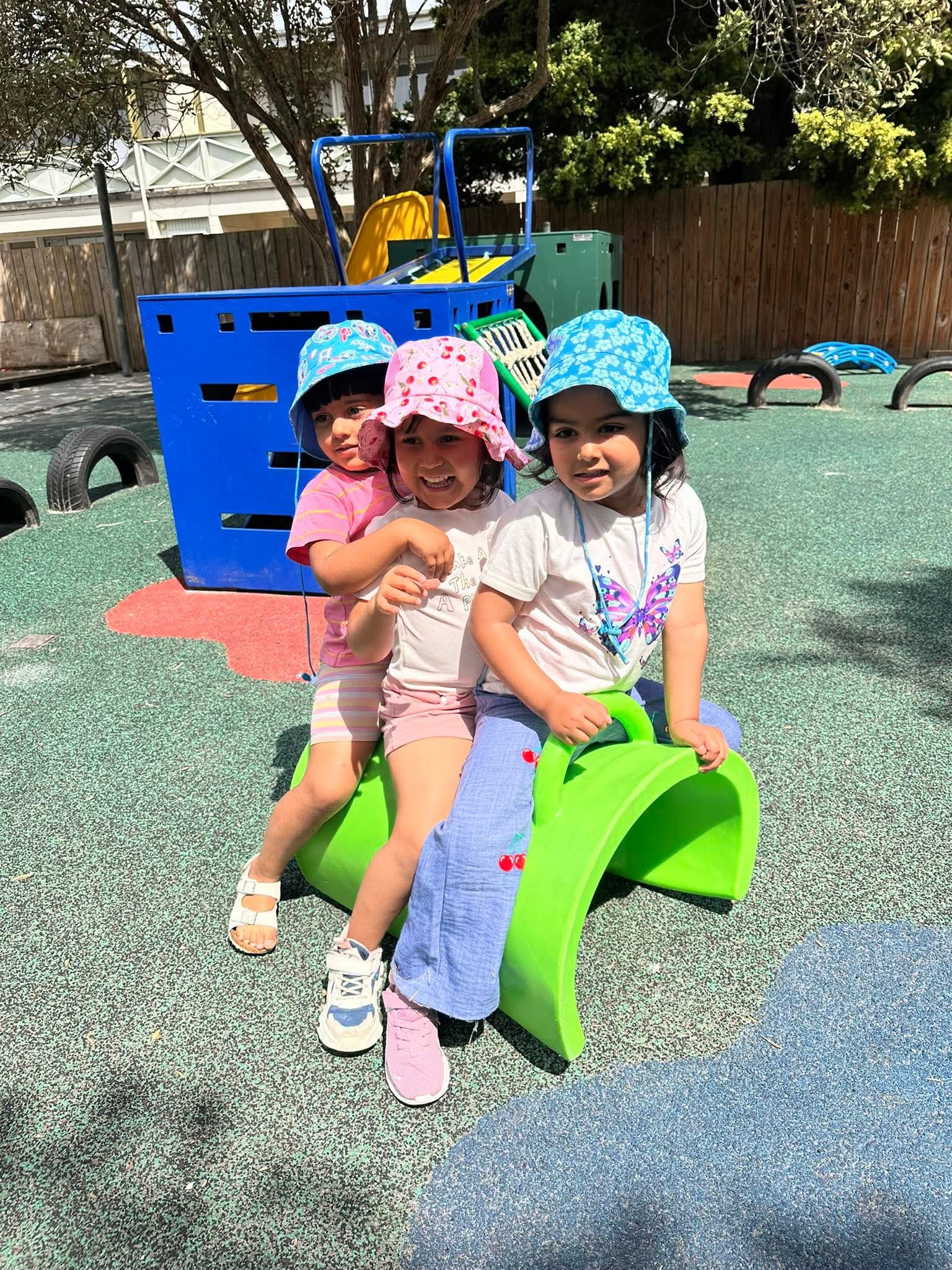 Three young children playing on a green plastic seesaw at a playground, wearing colorful sun hats and casual clothes, with playground equipment and a wooden fence in the background.