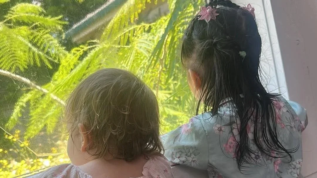 Two children with dark, wet hair looking out of a window at lush green plants and foliage outside.