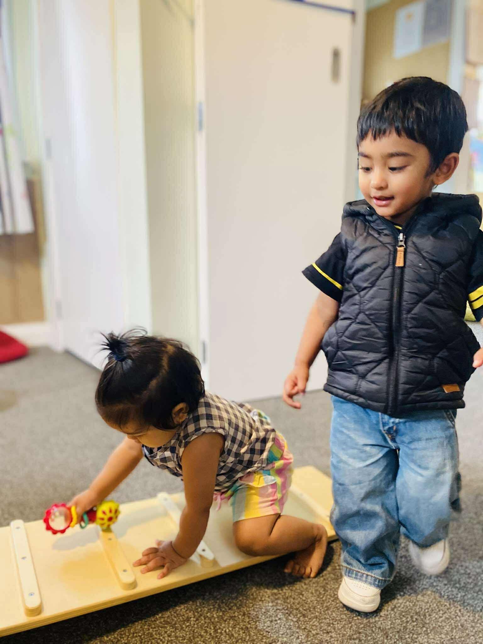 A young girl crouching on the floor playing with a toy car, while an older boy stands nearby watching, in an indoor setting.