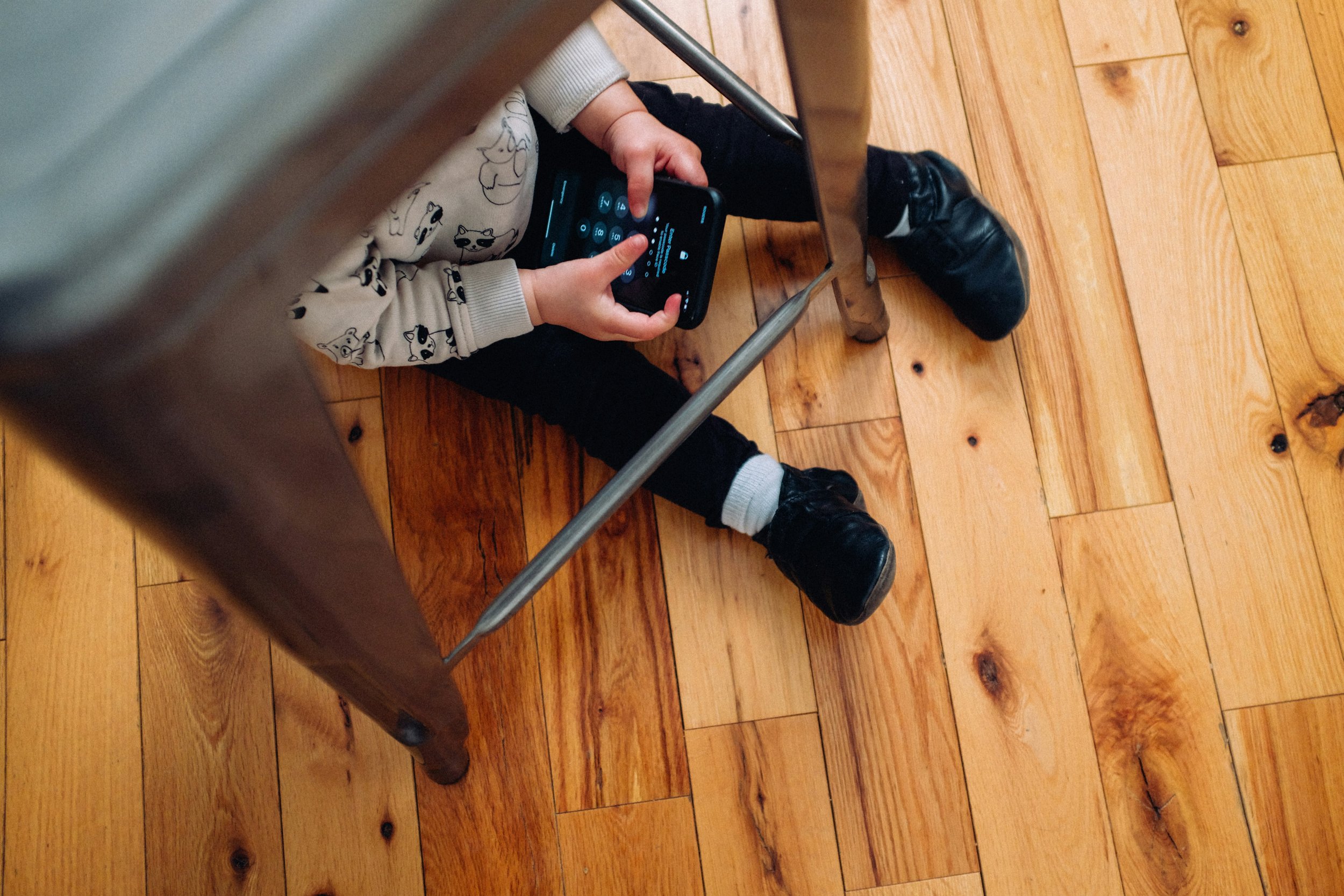 Child sitting on a hardwood floor underneath a table, holding a smartphone with one hand and using the other to tap the screen.