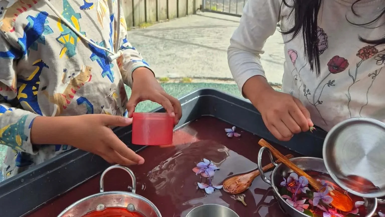 Two people pouring and handling purple flowers in a container on a dark red liquid surface outdoors.