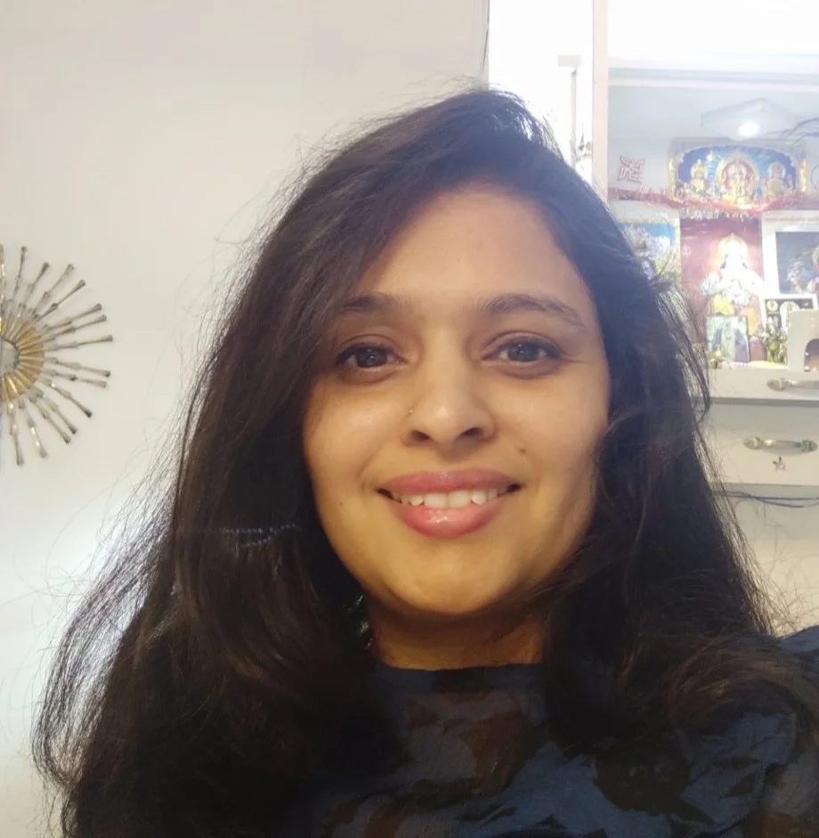 Close-up of a woman with dark hair, smiling, in a room with Hindu religious decorations and artwork in the background.