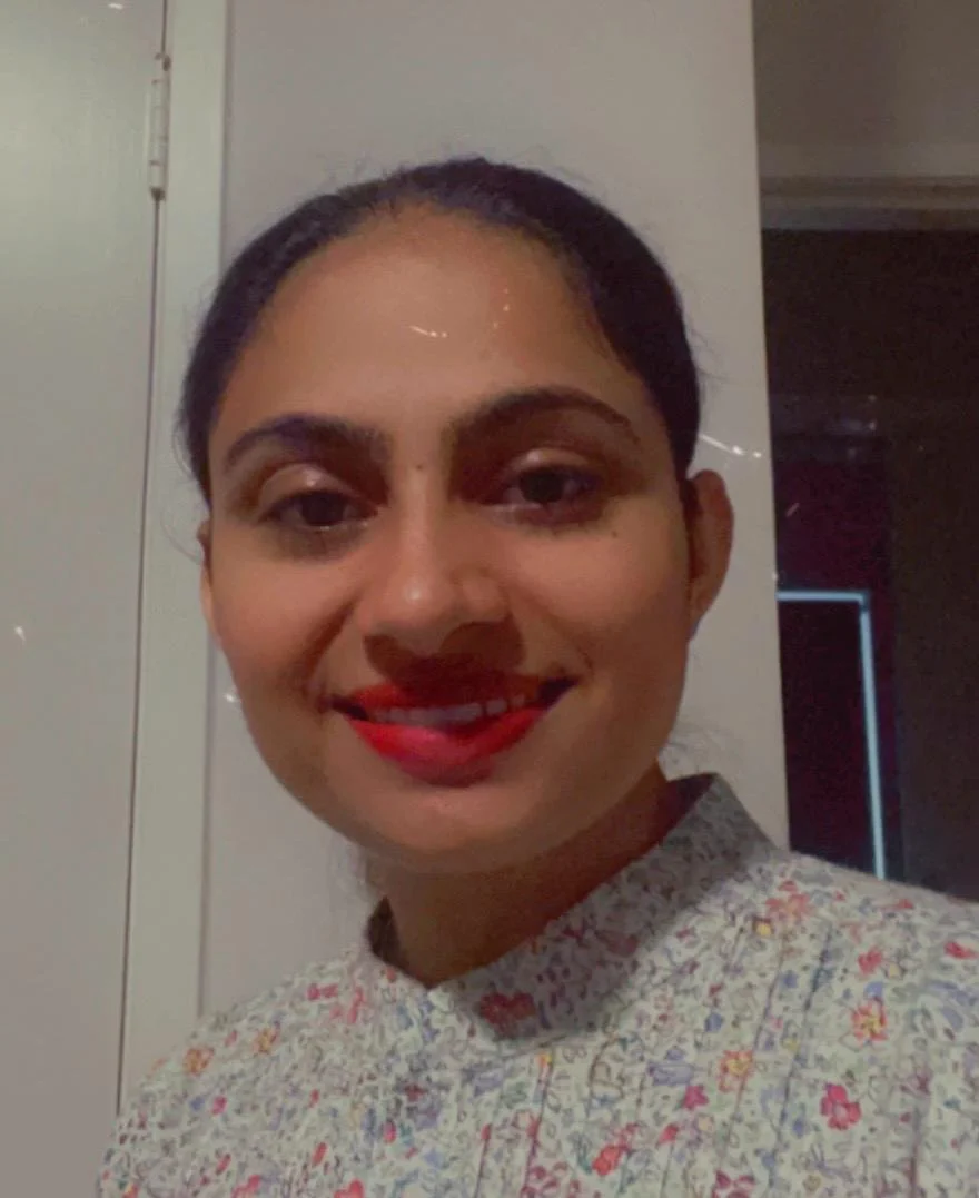 A woman with dark hair tied back, smiling, wearing a light-colored shirt with a floral pattern, standing indoors against a light-colored wall.