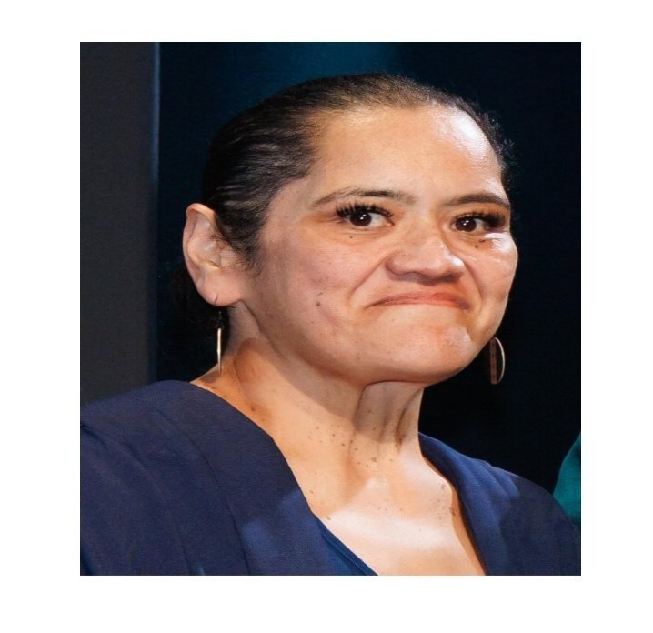 Close-up photograph of a woman with dark hair, wearing hoop earrings, and a navy blue top, making a facial expression with lips pressed together and slightly raised eyebrows, against a dark background.