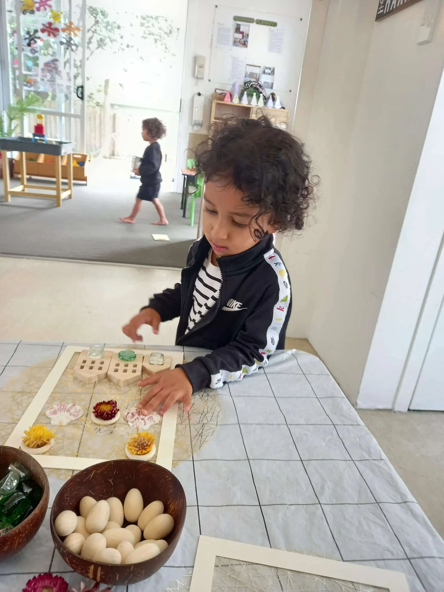 A young child with curly hair and a striped shirt, wearing a black jacket, is arranging small decorative items on a table in a classroom. Two bowls, one filled with white eggs and the other with small green objects, are on the table. In the background, another child walks in a room with natural light and decorations.