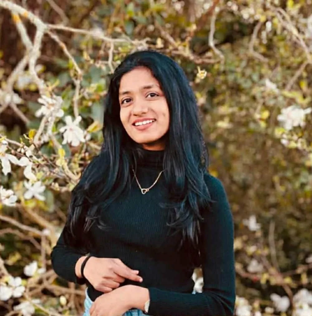 A young woman with long black hair and a black turtleneck sweater standing outdoors in front of blossoming trees with white flowers.