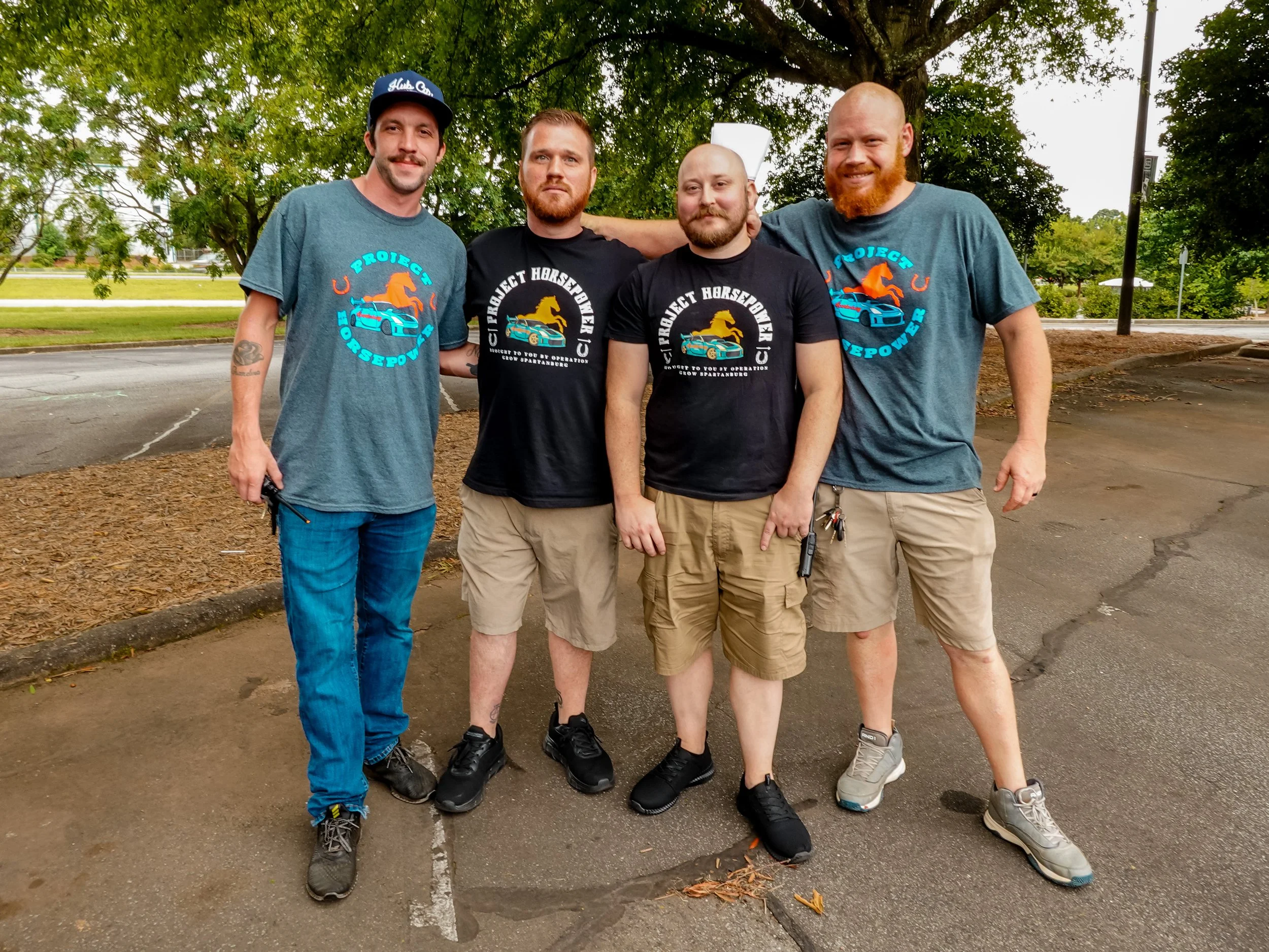 Five men standing outdoors under a tree, posing for a photo. They are wearing casual clothing and matching t-shirts with a racing car and horse logo, some with black shirts and some with gray. The background includes a parking lot, some grass, and trees.