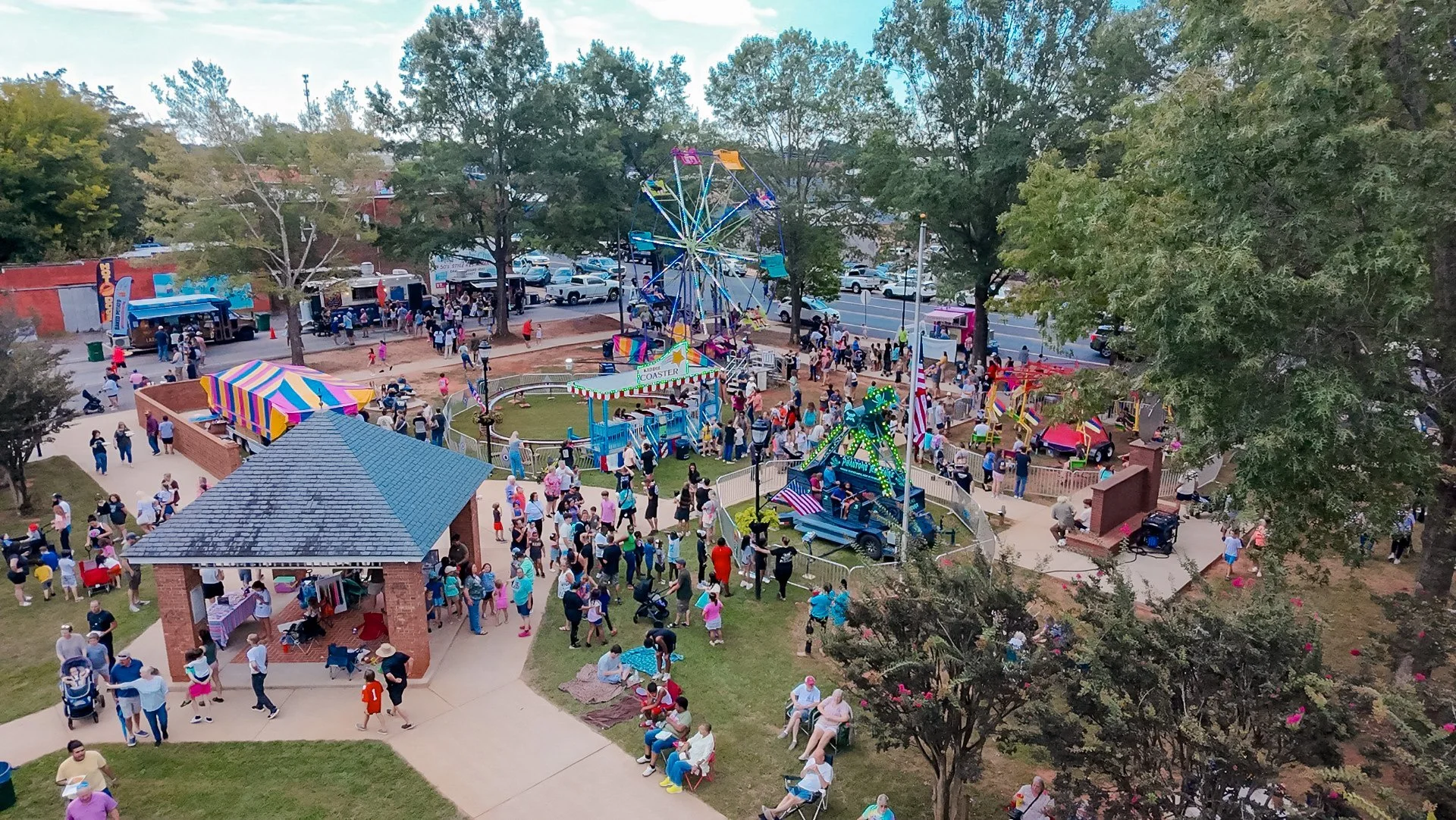A lively outdoor amusement park with a ferris wheel, a kiddie coaster, a carousel, and other rides. People are walking around, waiting in line, and sitting on benches. There are food stands and trees surrounding the park.