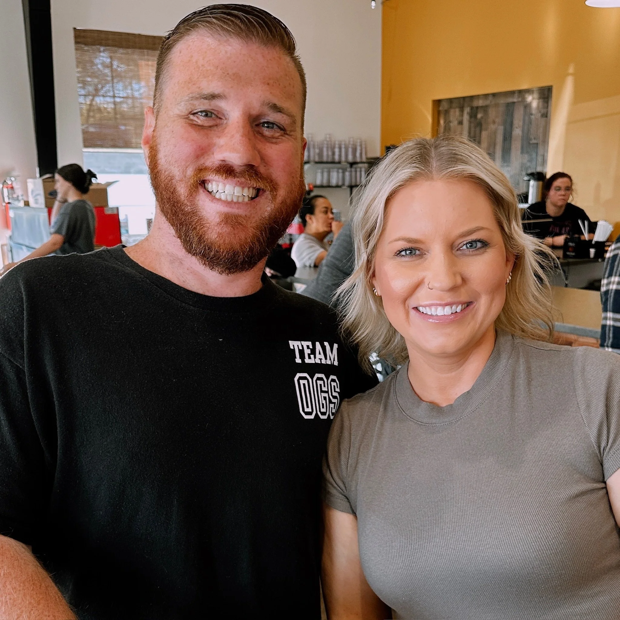 A man and woman smiling at the camera in a cafe or restaurant with other people in the background.