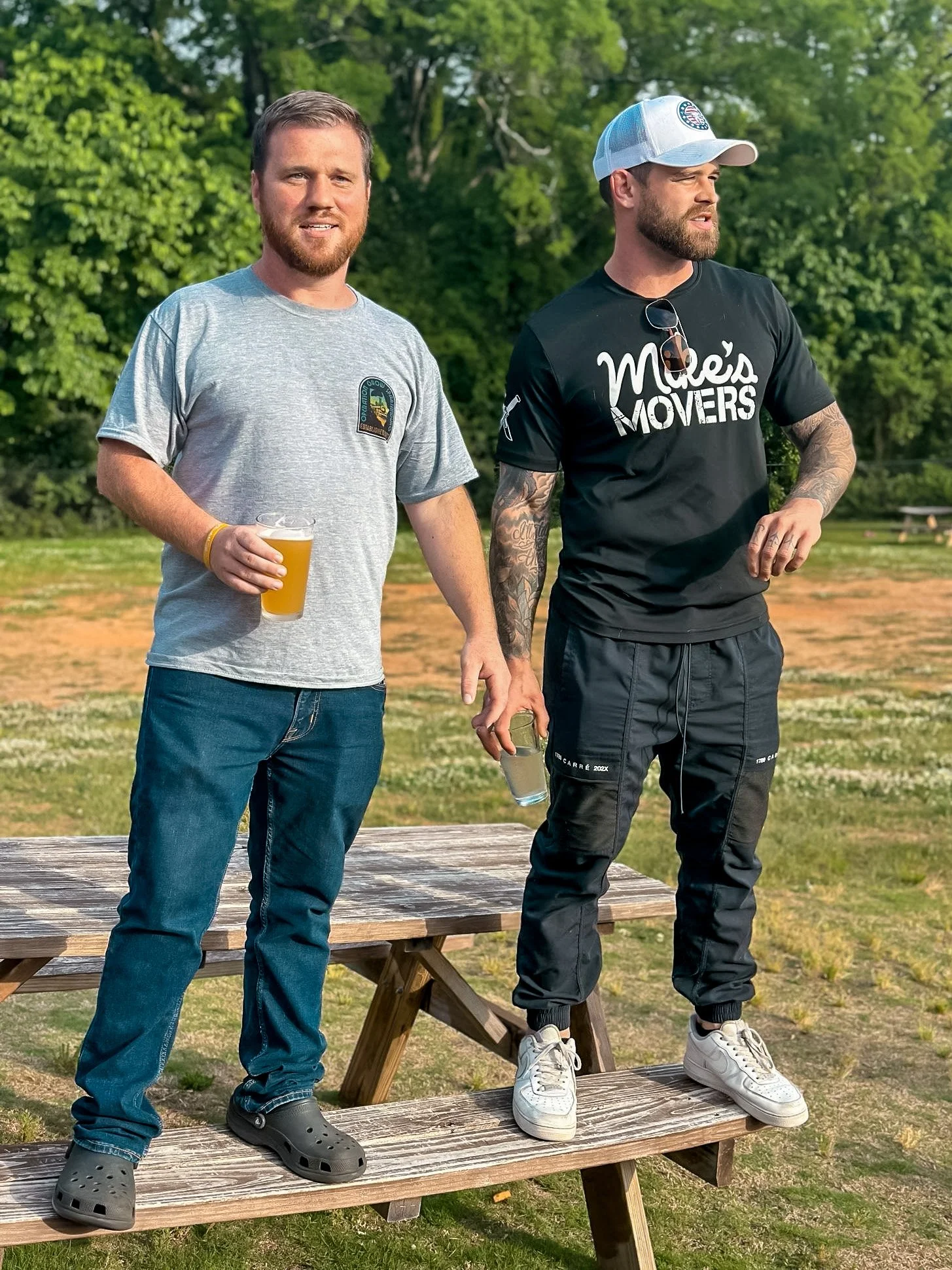 Two men standing on outdoor picnic tables holding drinks with green trees in the background.