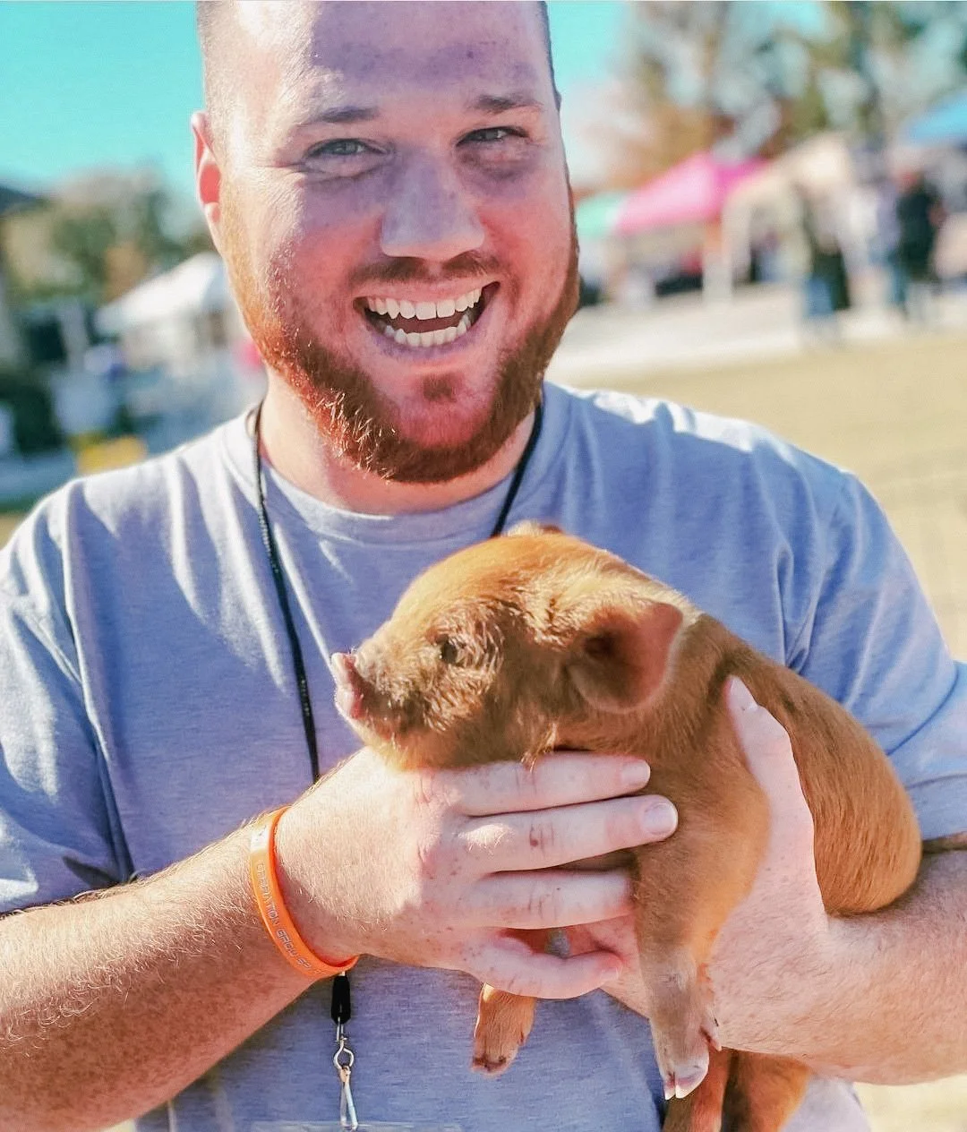 A man with a beard smiling and holding a small brown animal, likely a piglet, outdoors at a fair or festival.