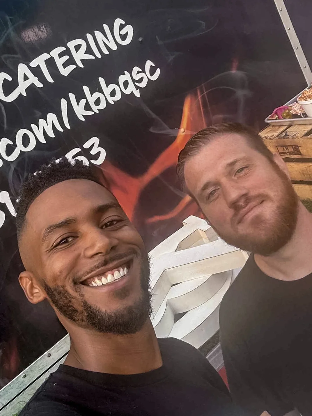 Two men smiling and posing for a selfie in front of a blackboard with white writing about catering, with plates of food visible on a table in the background.