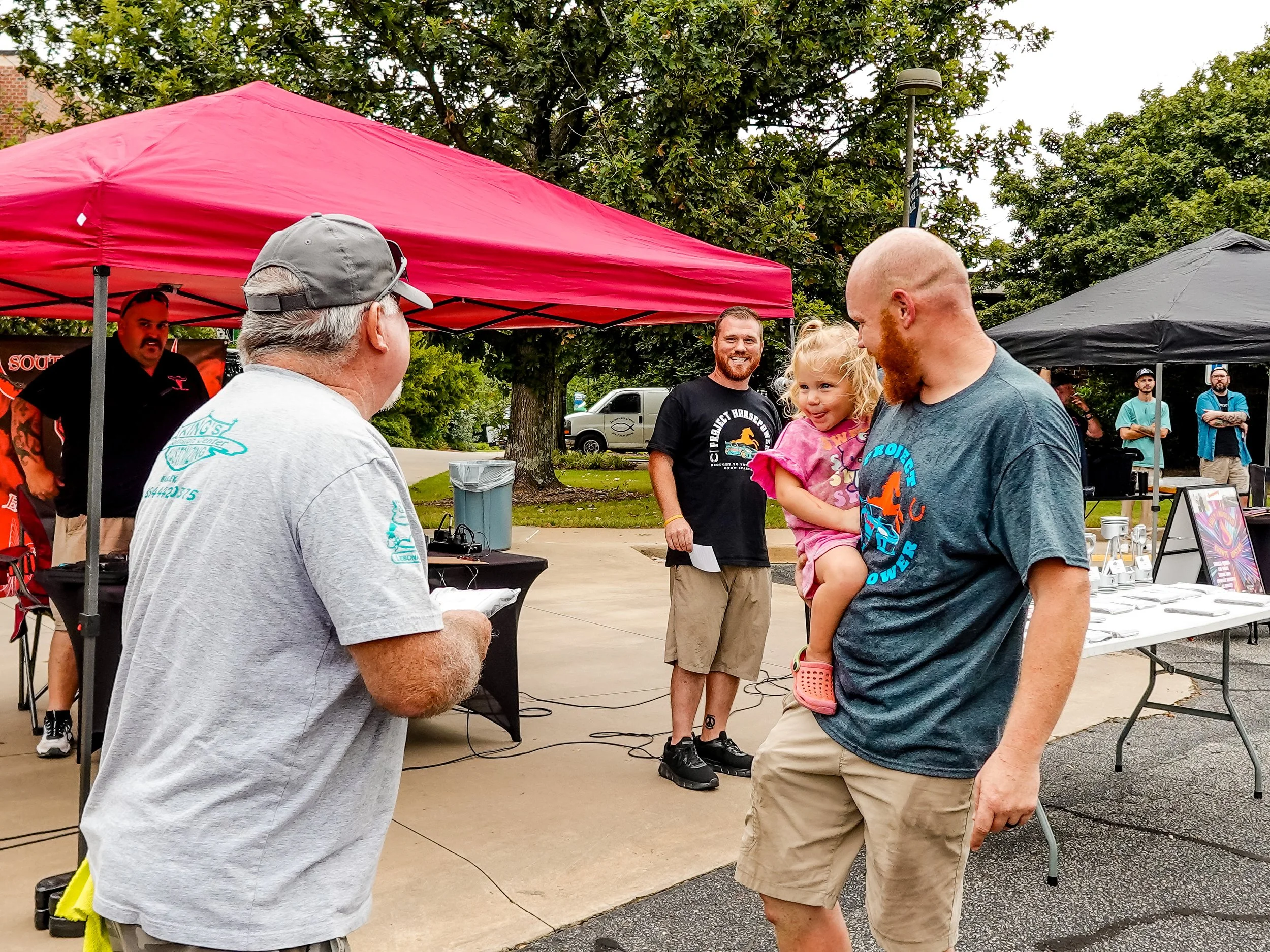 A man with a beard and gray T-shirt holding a young girl in pink, standing outdoors at a community event. Other people are present, with a red canopy canopy and a table with items in the background.