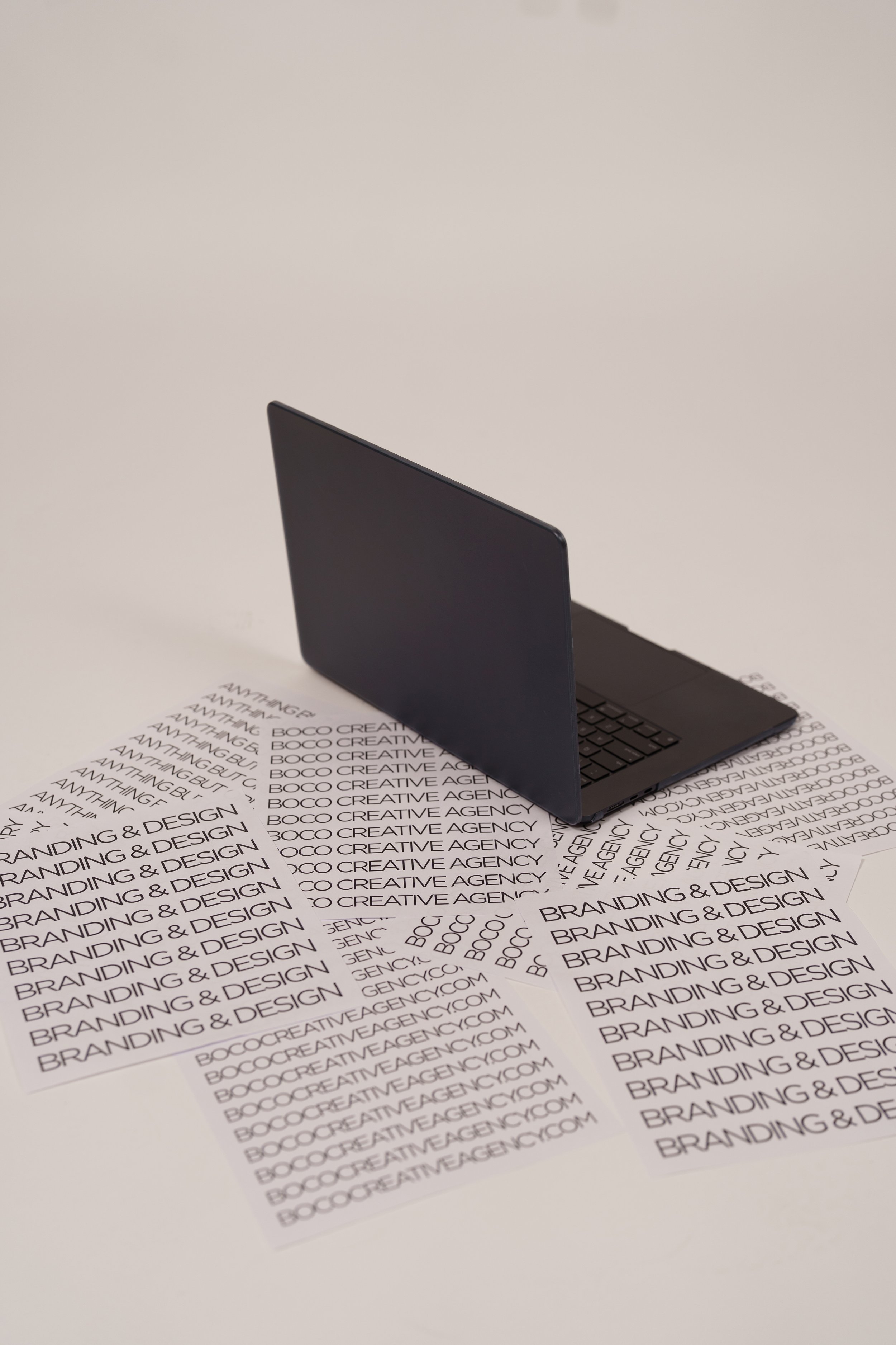 A closed black laptop on a white surface with sheets of paper printed with "BRANDING & DESIGN" and "BOCCREATIVEAGENCY.COM" in black text.